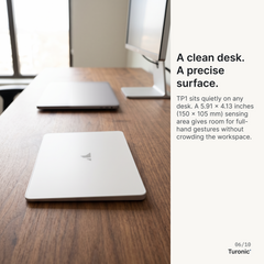 Silver rectangular touchpad with Turonic logo resting flat on a wood desk, with a laptop and monitor visible behind it.