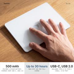 A hand with four fingers resting flat on the white glass touchpad sitting on a wooden desk.